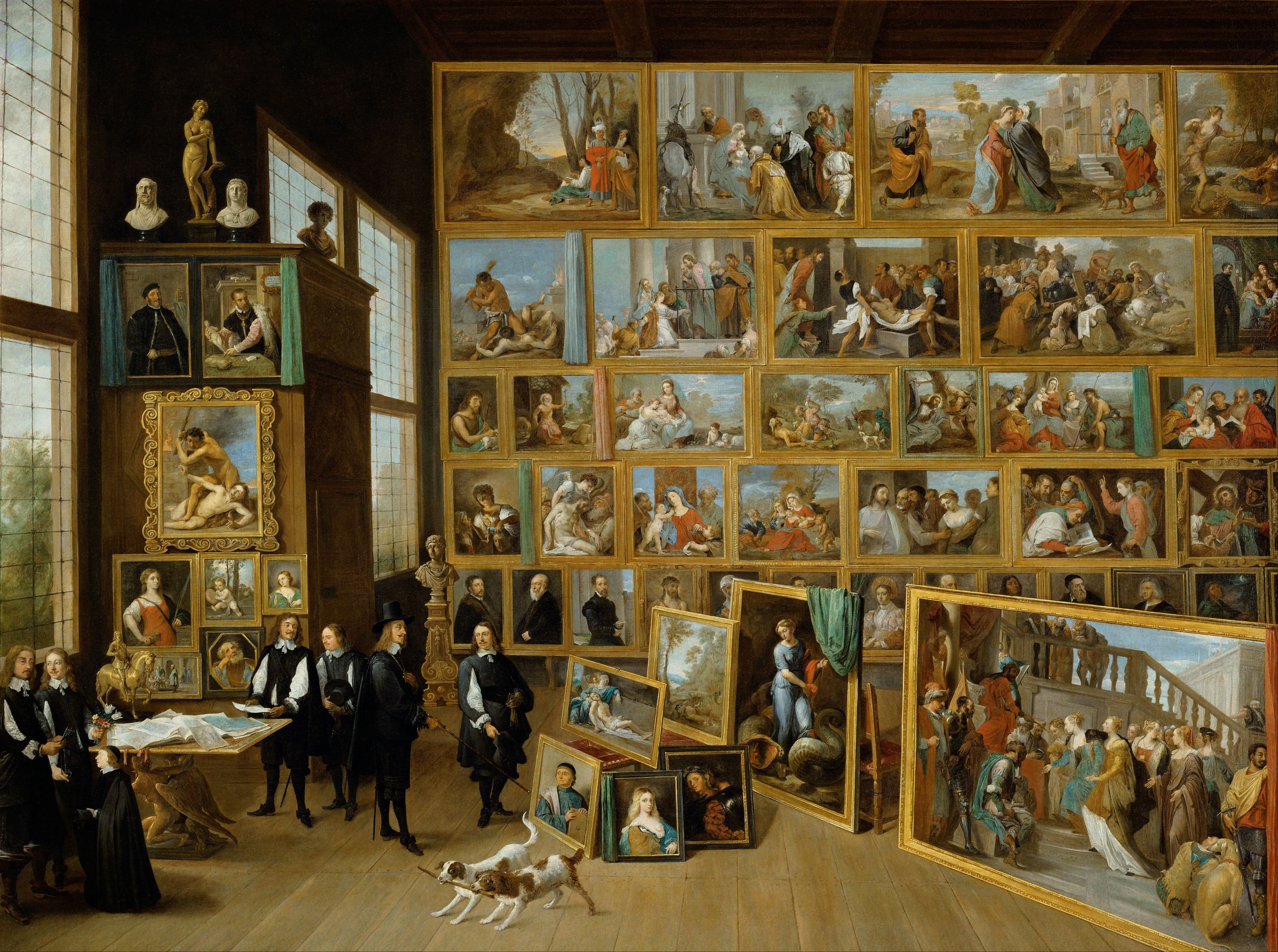 David Teniers’s painting of the Archduke’s picture gallery: tall windows on the left, a wall covered in gilt-framed paintings on the right, men in black at the center, two dogs, and new paintings leaning on the floor.