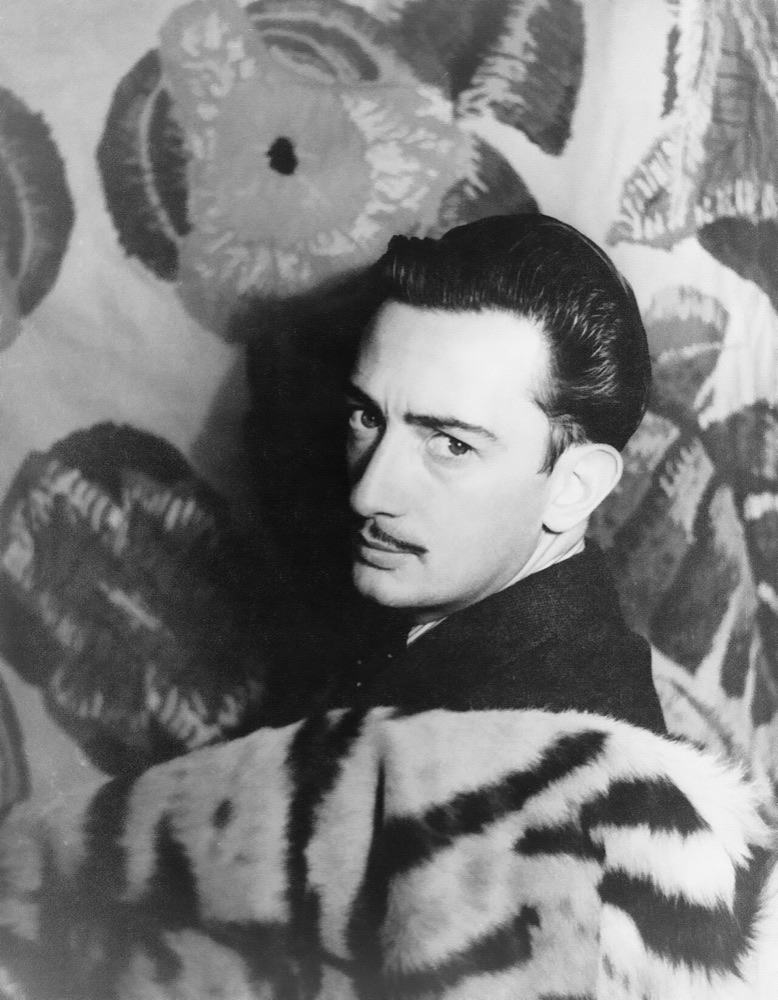 A black-and-white studio portrait of Salvador Dalí in 1939, glancing sideways at the camera with a thin mustache, in front of a wall hanging of large flowers.