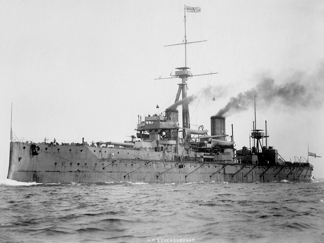 A black-and-white photograph of the battleship HMS Dreadnought under way, about 1906, with two funnels, a tall tripod mast and smoke trailing behind.