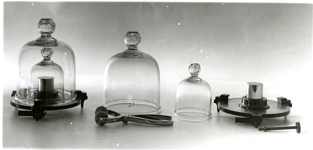A black-and-white photograph of two small metal cylinders, one sealed under a double glass bell jar and one standing uncovered beside its two bell jars.
