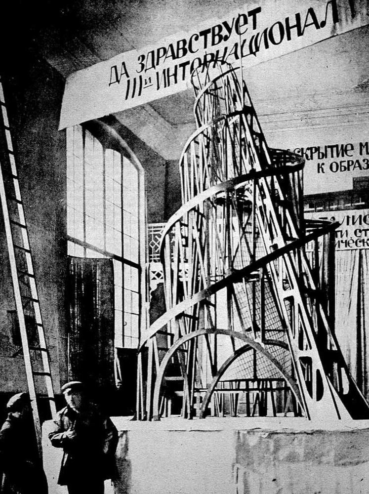 Black and white photograph of the wooden model of Tatlin’s tower in a Petrograd hall in 1920: a leaning double spiral of lattice beams over two arches, with a banner in Russian overhead and two men standing at its foot.